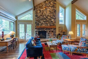Amplia sala de estar con chimenea de piedra, ventanas grandes y una mezcla de muebles modernos y tradicionales.
