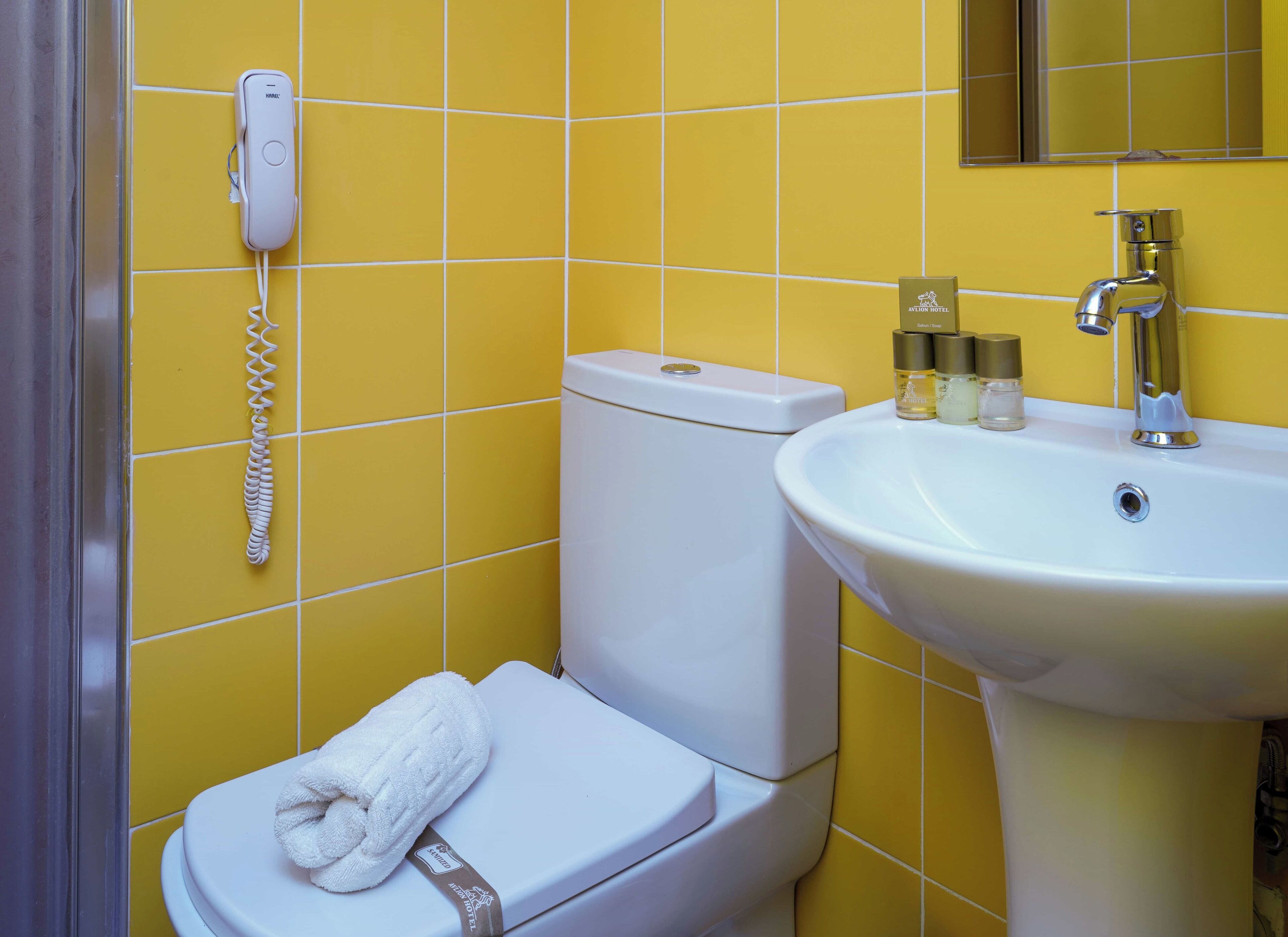 Une salle de bain avec un mur recouvert de carreaux jaunes, des toilettes blanches, un lavabo blanc avec des articles de toilette et un téléphone fixé au mur.
