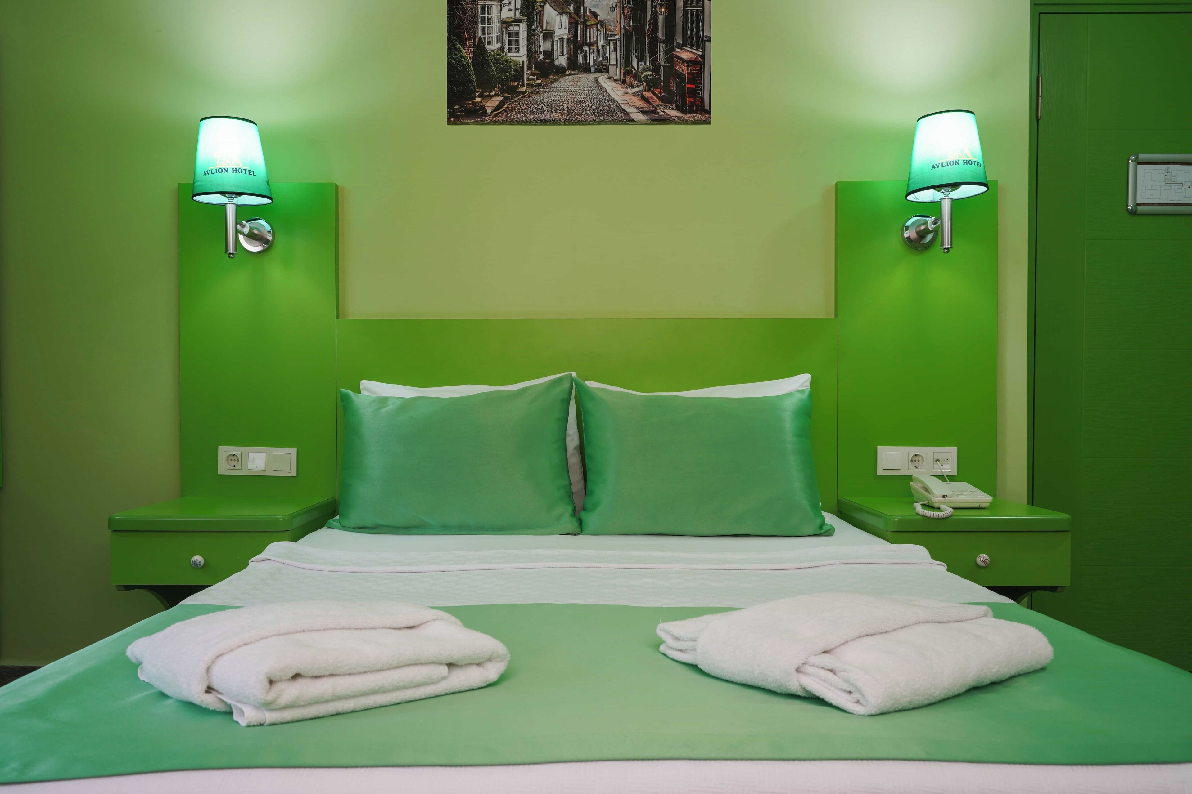 Habitación de hotel con decoración verde, que incluye una cama, dos mesitas de noche verdes, un teléfono y dos lámparas de pared.