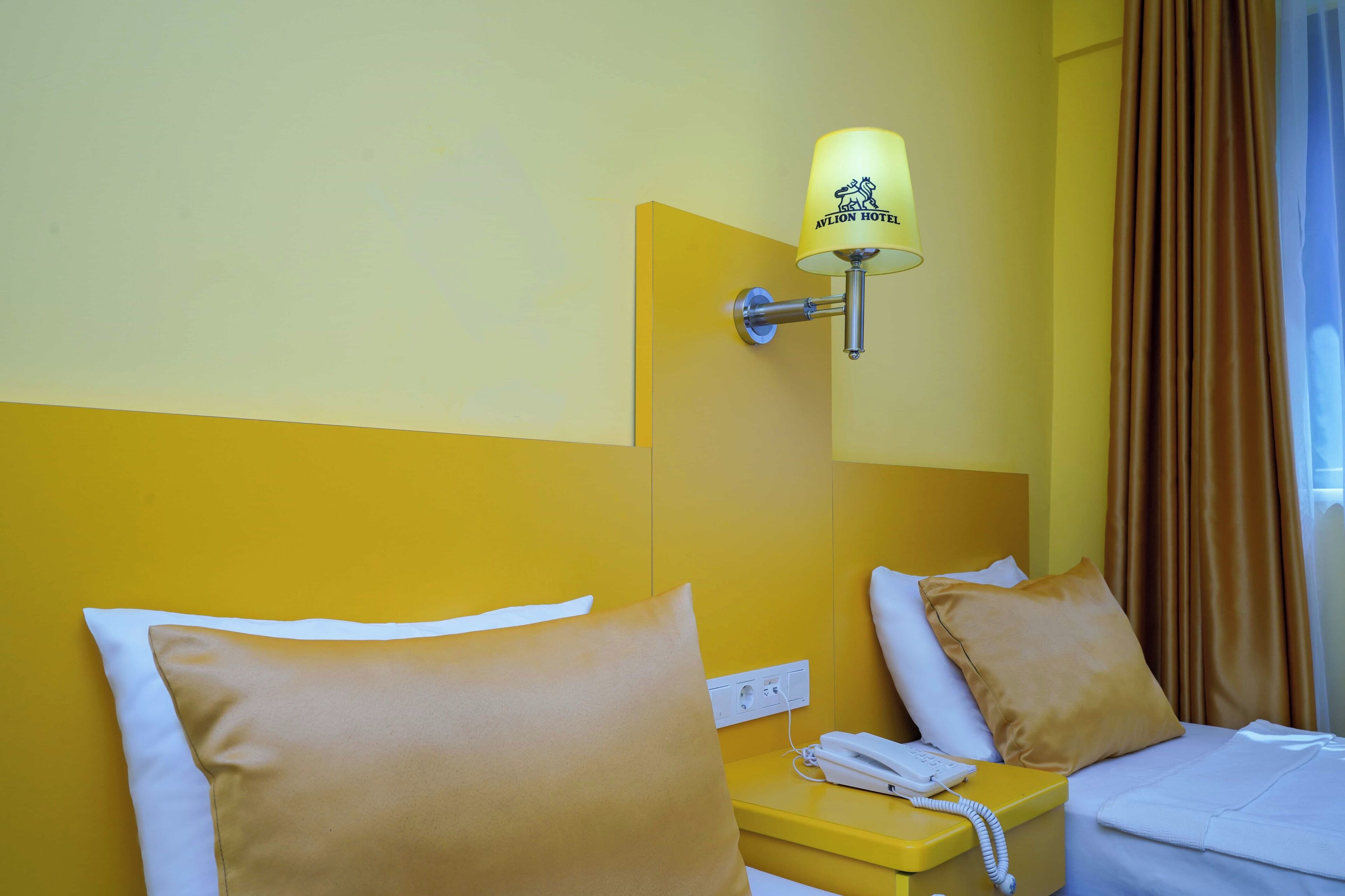 A hotel room with a bed, a bedside table with a phone, a lamp, and a power outlet.