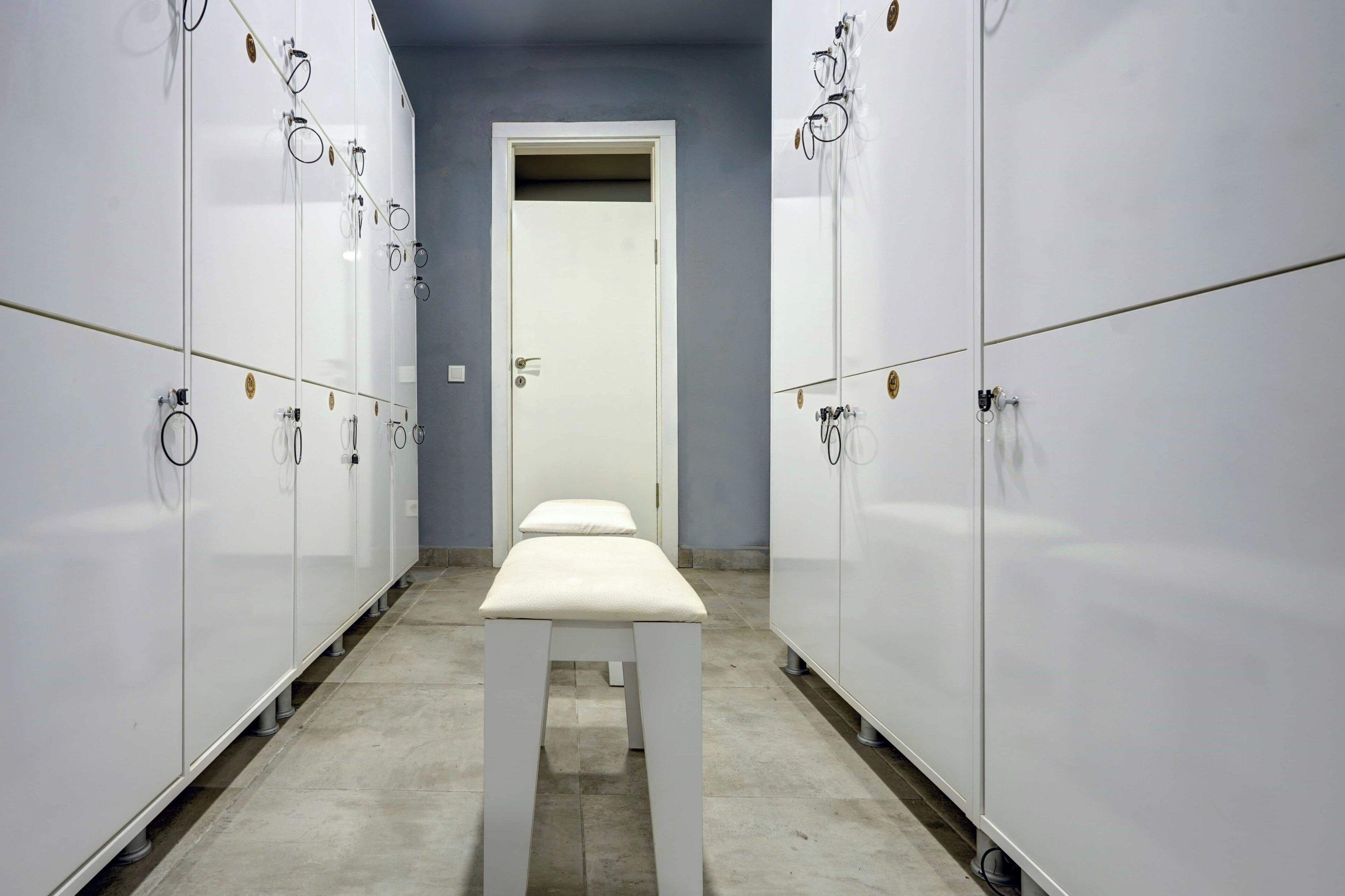 A corridor with white lockers on both sides, a bench in the center, and a door at the end.
