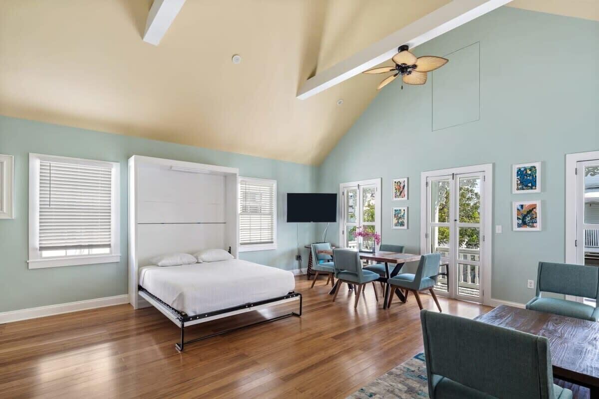 A bedroom with a bed, a desk, and chairs, a ceiling fan, and a TV.