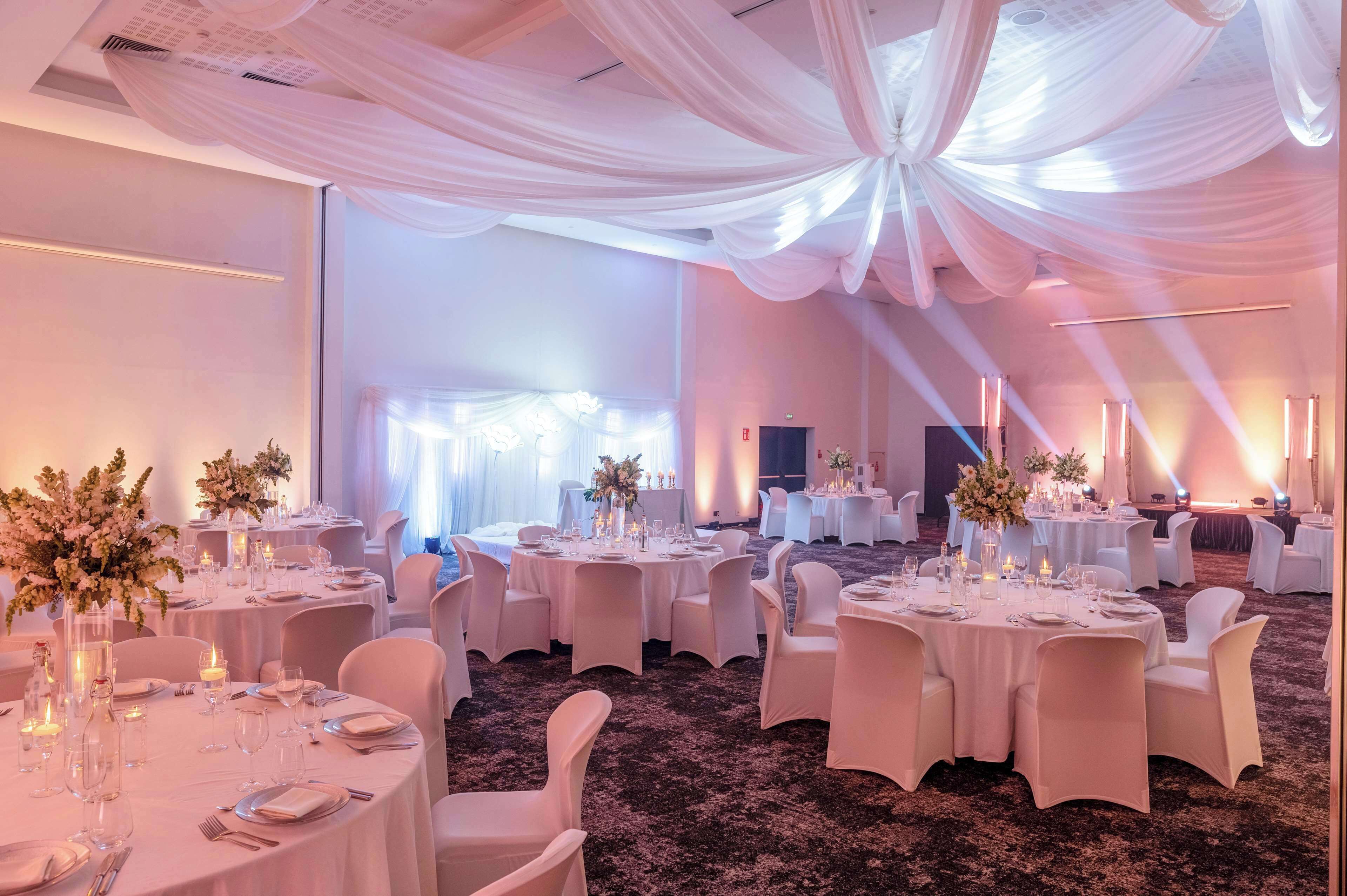 A banquet hall with round tables set for dining, white drapery on the ceiling, and a carpeted floor.