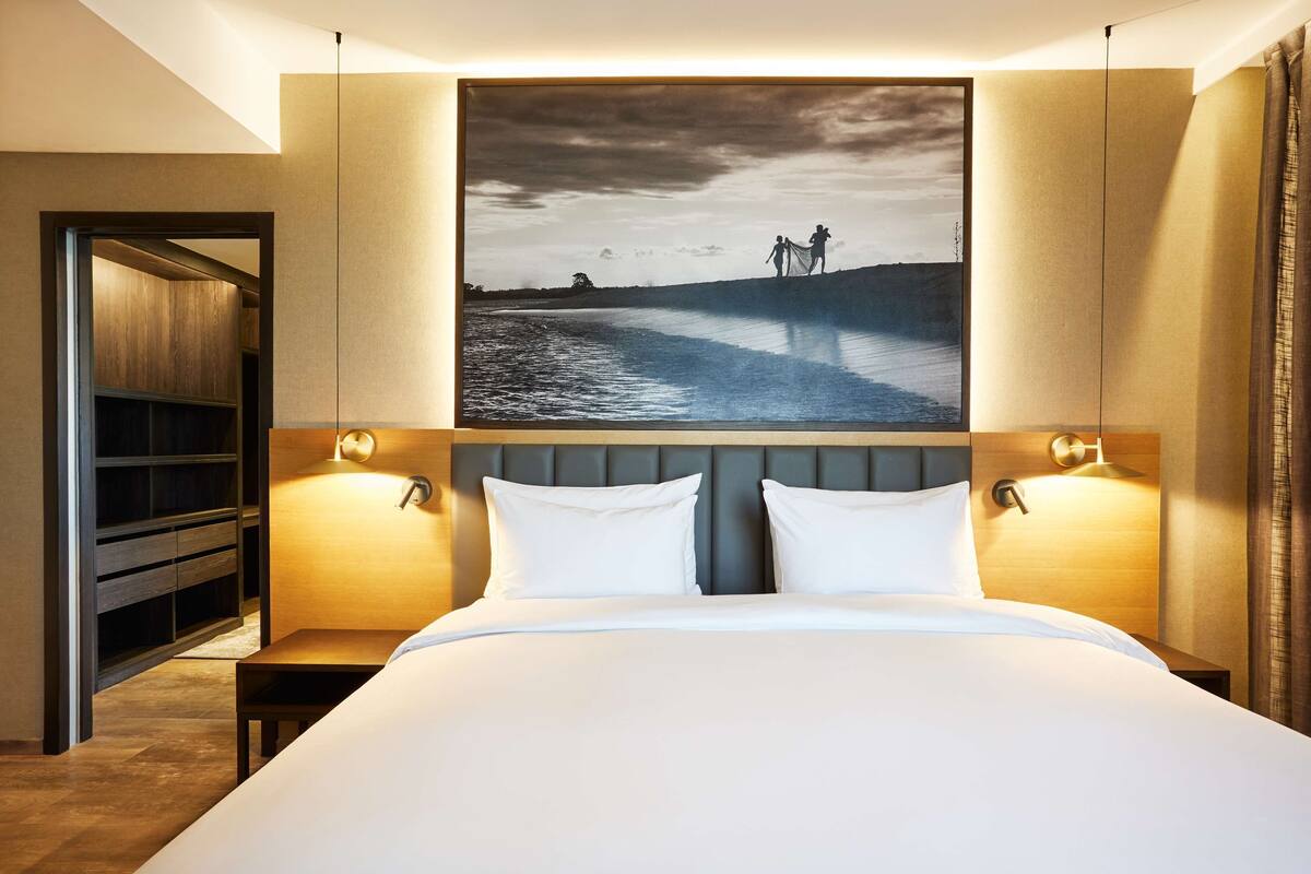 Habitación de hotel con una cama grande, dos mesitas de noche, un cabecero y un cuadro colgado en la pared que muestra un paisaje.