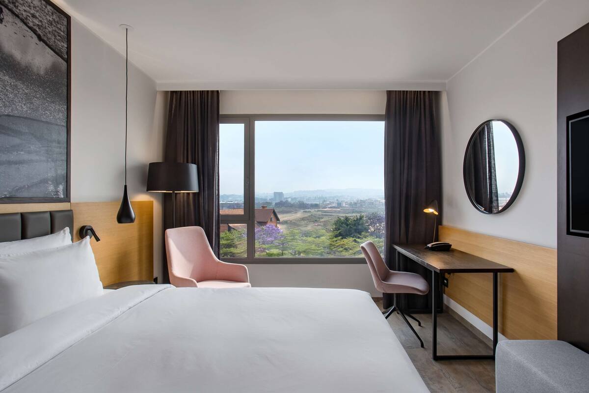 Habitación de hotel con una cama grande, un escritorio, dos sillas, vistas de la ciudad y un espejo redondo en la pared.
