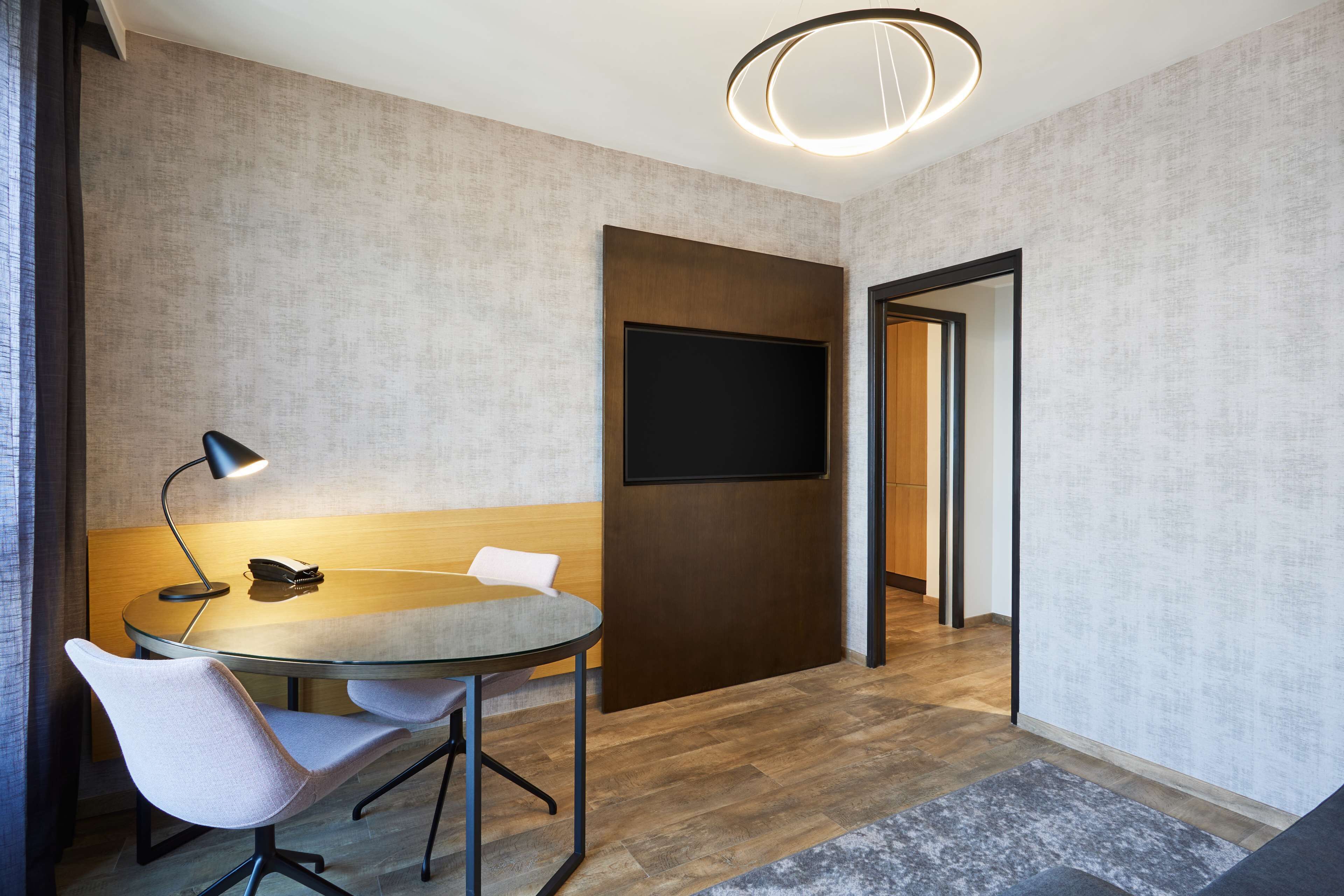 A modern conference room with a glass table, two chairs, a desk lamp, a mounted TV, and a wooden accent wall.