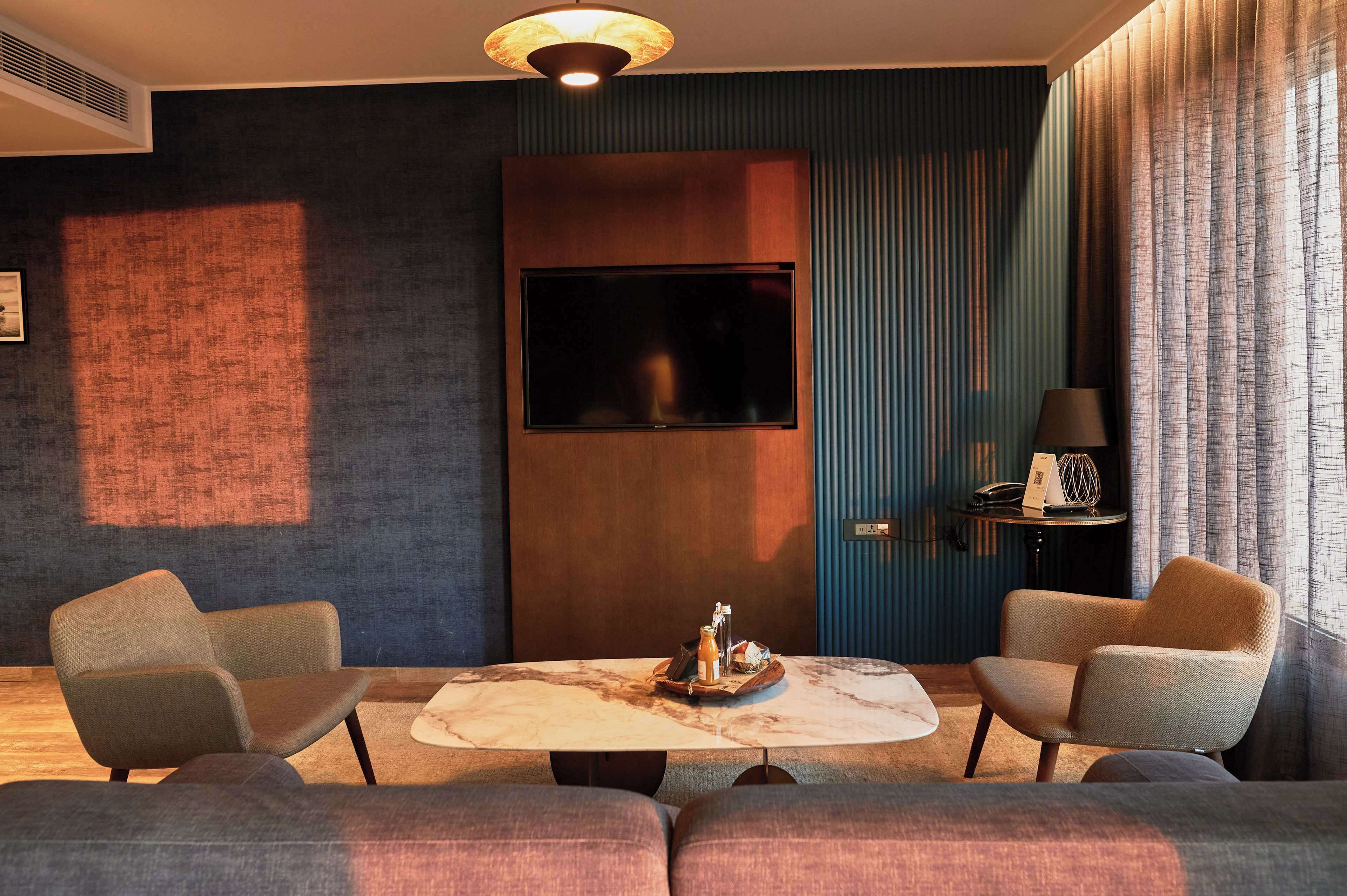 A modern hotel room with a large flat-screen TV mounted on the wall, a marble coffee table with a tray of items, two armchairs, and a side table with a lamp.