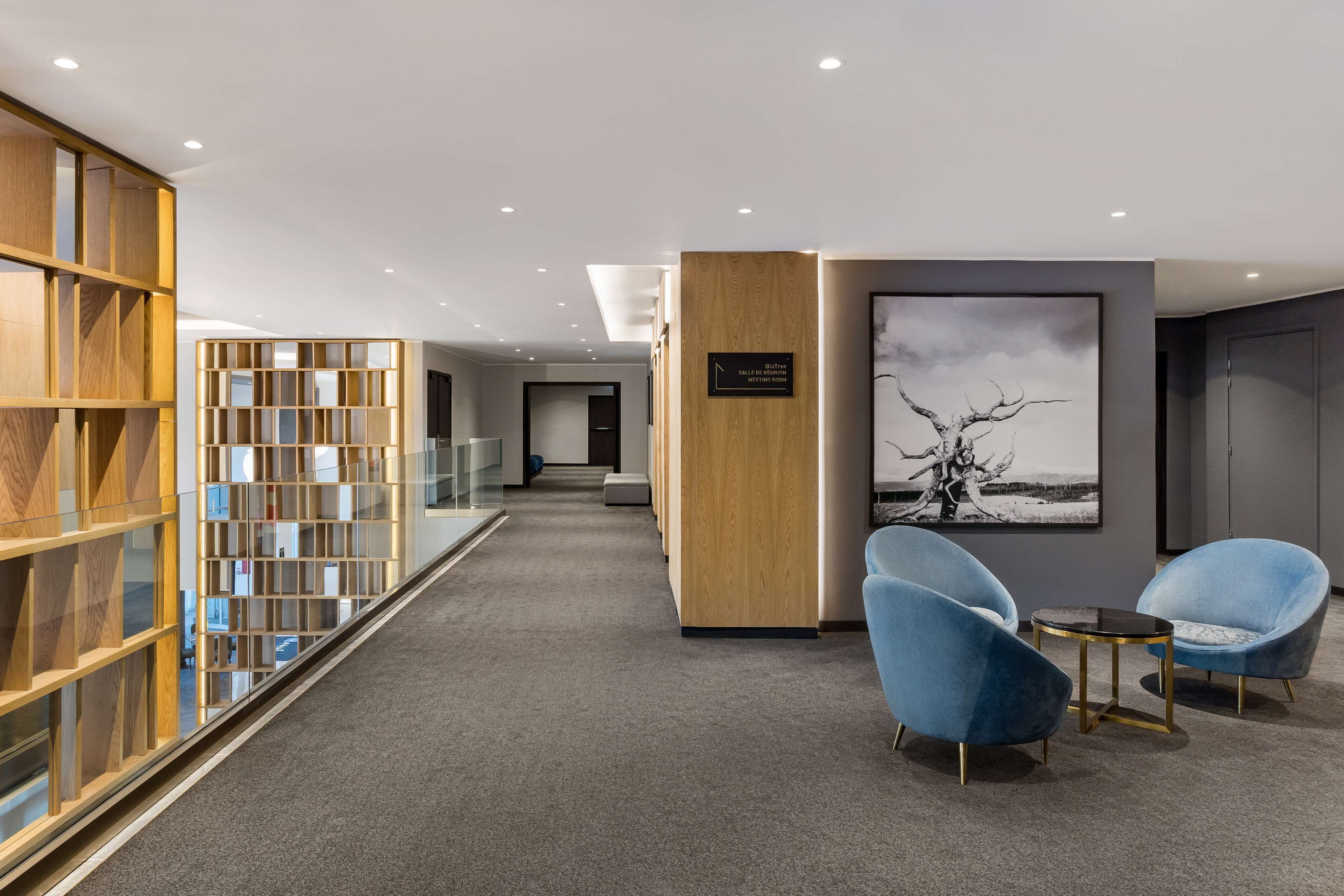 Un moderno hall de hotel con un mostrador de recepción, una zona de estar con sillas azules, una estantería y una obra de arte enmarcada en la pared.