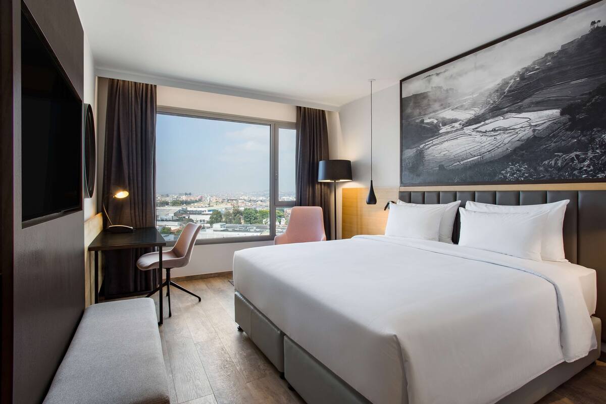 Habitación de hotel con una cama grande, un escritorio, una silla, un televisor y una ventana con vistas a la ciudad.