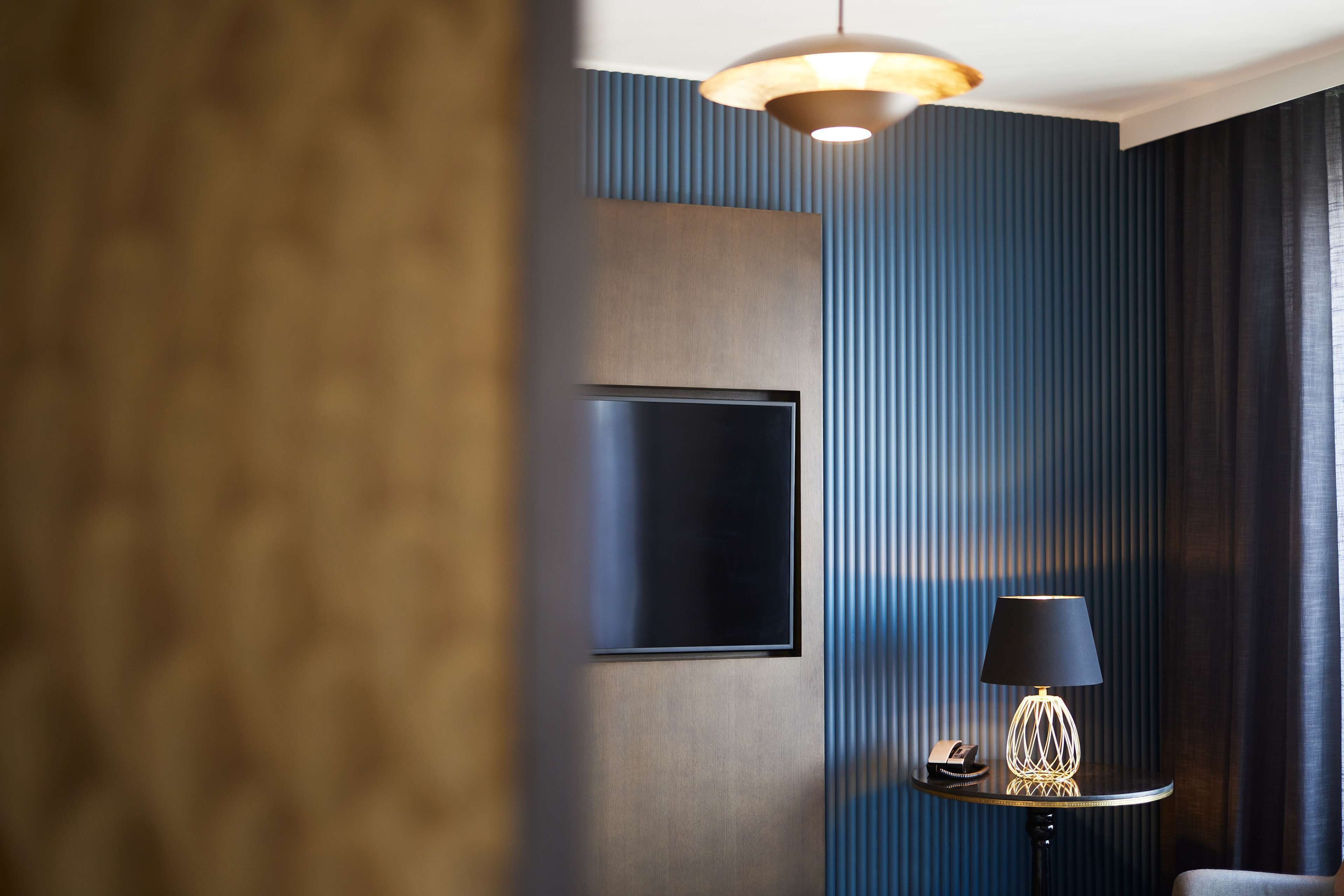 A modern hotel room with a large flat-screen TV mounted on the wall, a round table with a lamp, and a telephone.