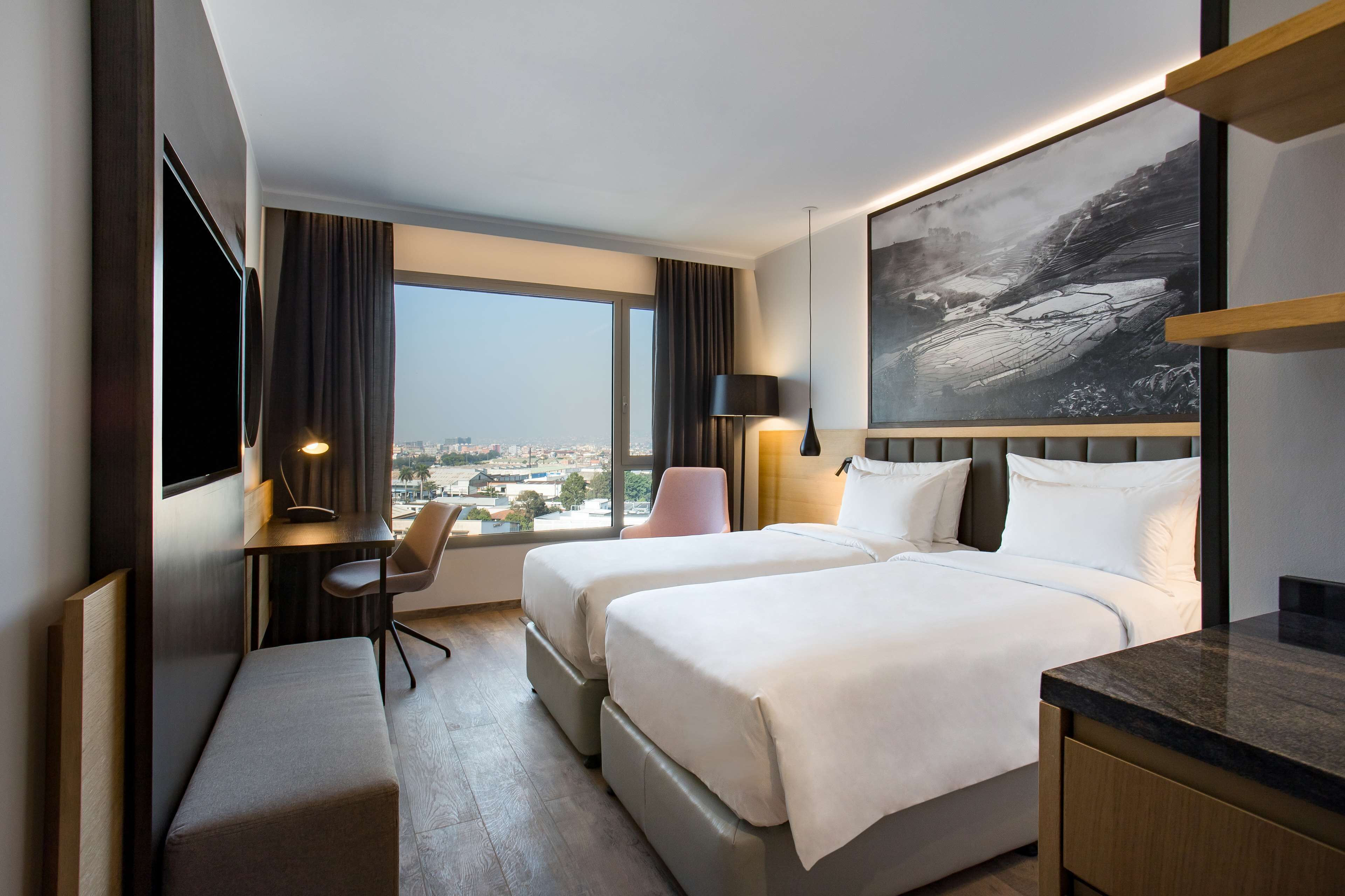 A hotel room with two beds, a desk, a chair, a bench, a TV, and a large window offering a city view.