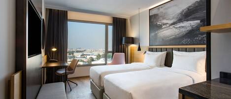 A hotel room with two beds, a desk, a chair, a bench, a TV, and a large window offering a city view.
