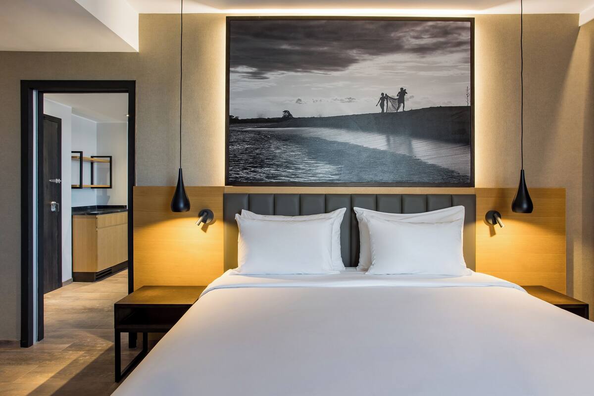 Una habitación de hotel moderna con una cama grande, dos lámparas de noche, una mesita de noche y un cuadro grande enmarcado sobre la cama.