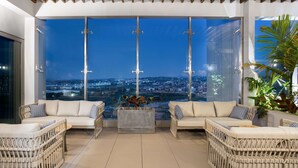 Un salón moderno con mobiliario de mimbre, un ventanal con vistas a la ciudad y un techo con vigas de madera.