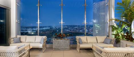 A modern lounge with wicker furniture, a large window offering a city view, and a ceiling with wooden beams.