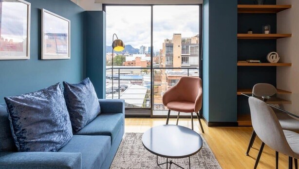 Una sala de estar moderna con un sofá azul, una mesa de centro redonda y una silla rosa. Hay un ventanal con vistas a la ciudad, y una estantería con algunos objetos.
