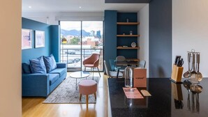 Una sala de estar moderna con un sofá azul, un sillón rosa, una mesa de centro y una zona de cocina con utensilios.