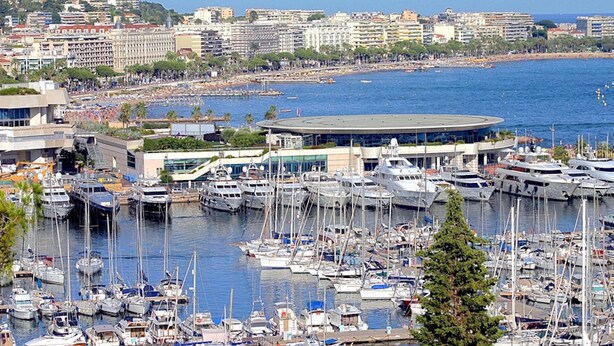 Una marina con numerose imbarcazioni ormeggiate, un ampio edificio circolare e uno skyline sullo sfondo.