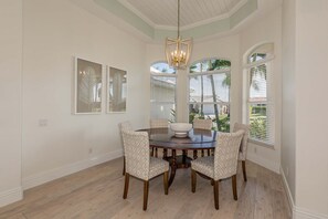 Un coin repas comprenant une table ronde, quatre chaises à motifs, un luminaire central et de grandes fenêtres équipées de stores.