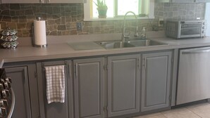 A kitchen with grey cabinets, a stainless steel sink, a toaster, and a window with a view of greenery. - SKI, FISH, GOLF, RAFT, HIKE, HUNT, ATV ride, RELAX, 4 bdr home on wooded ADK lot (Malone)
