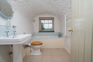 Une salle de bain avec un lavabo blanc, des toilettes en bois, une baignoire et une fenêtre avec des stores.