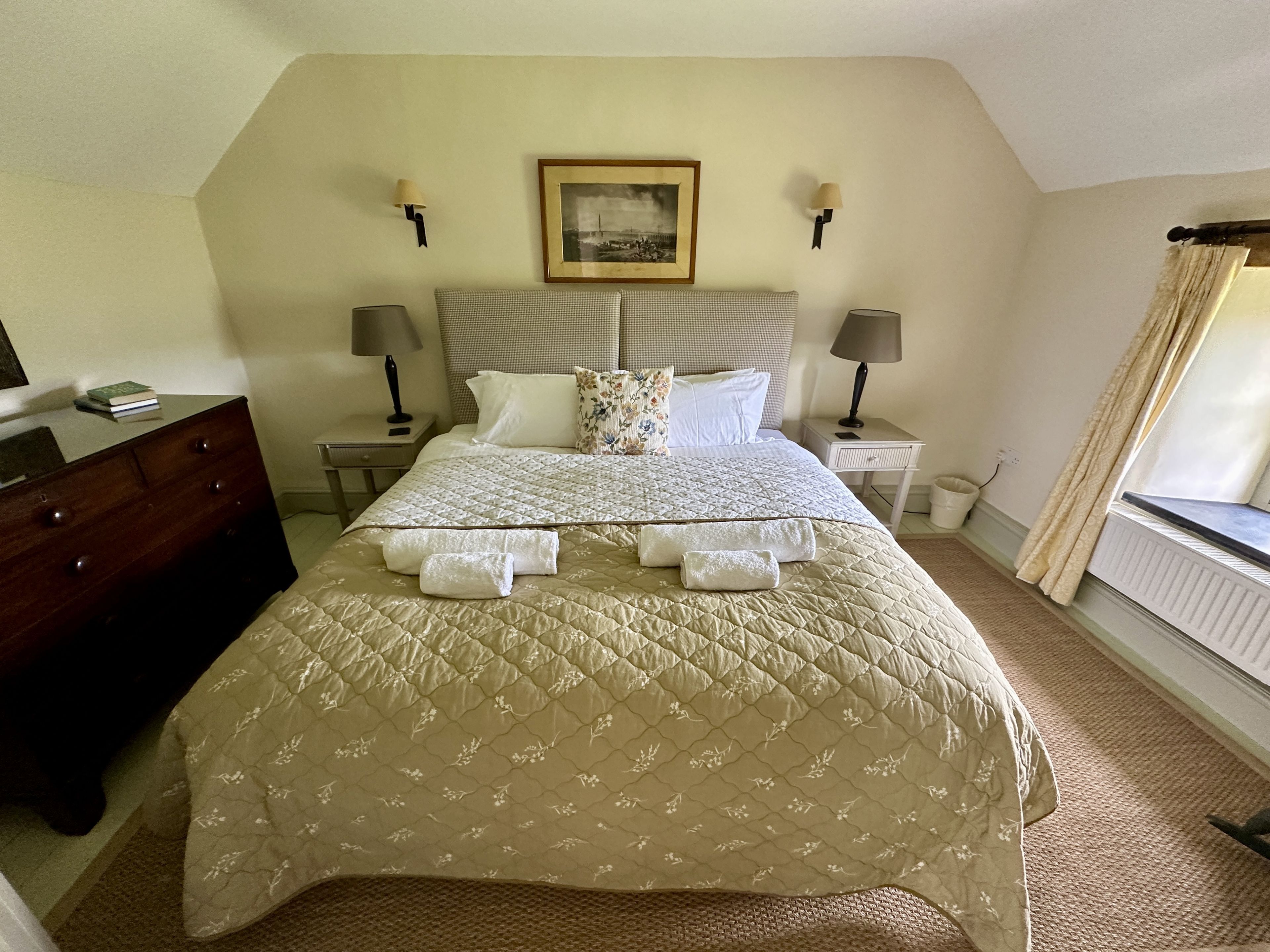 A neatly made bed with a quilted cover, two bedside lamps, a framed picture above the bed, and a dresser to the left.