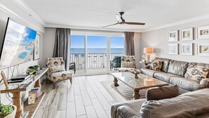 Amplia sala de estar con balcón, ventilador de techo y un gran ventanal con vista al mar.
