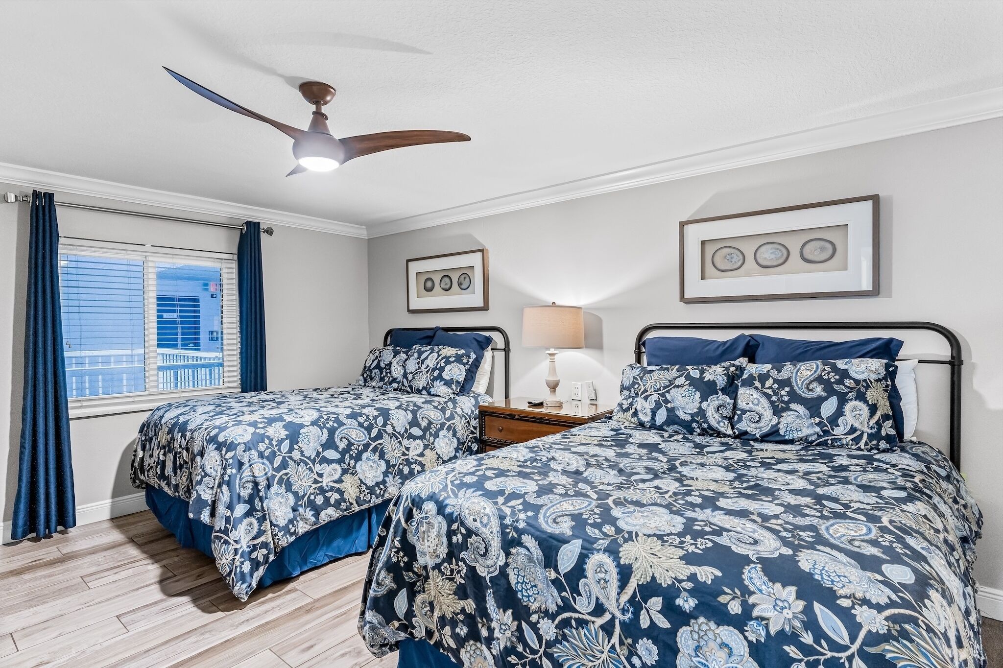 A bedroom with two beds, a ceiling fan, a window with curtains, and framed pictures on the wall.