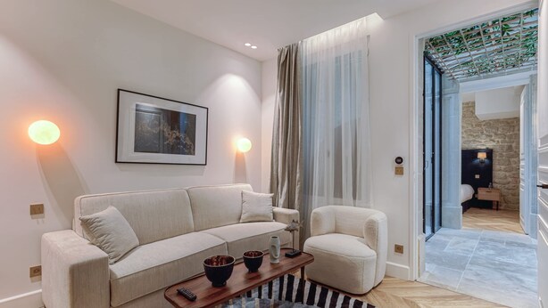 A modern living room with a sofa, armchair, and a coffee table. There is a framed picture on the wall, a window with curtains, and a view of a stone wall and a door leading to another room. - St Georges - Incredible One Bedroom with a patio (Paris)