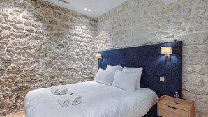 A bedroom with a stone wall, a bed with white linens, two bedside lamps, and a wooden nightstand with a bottle. - St Georges - Incredible One Bedroom with a patio (Paris)