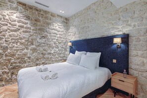A bedroom with a stone wall, a bed with white linens, two bedside lamps, and a wooden nightstand with a bottle. - St Georges - Incredible One Bedroom with a patio (Paris)