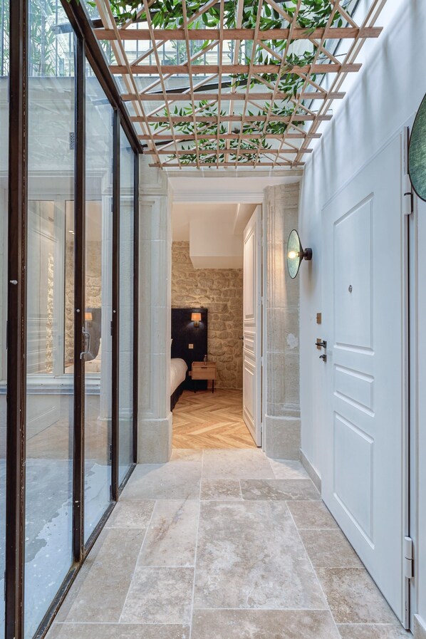 A hallway with a glass door leading to a bedroom, a wooden pergola with greenery, a stone wall, and a bed with a nightstand.