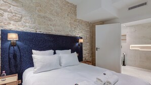 A hotel room with a stone wall, a bed with white bedding, two bedside lamps, and a wooden nightstand with a bottle.