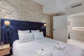A hotel room with a stone wall, a bed with white bedding, two bedside lamps, and a wooden nightstand with a bottle.