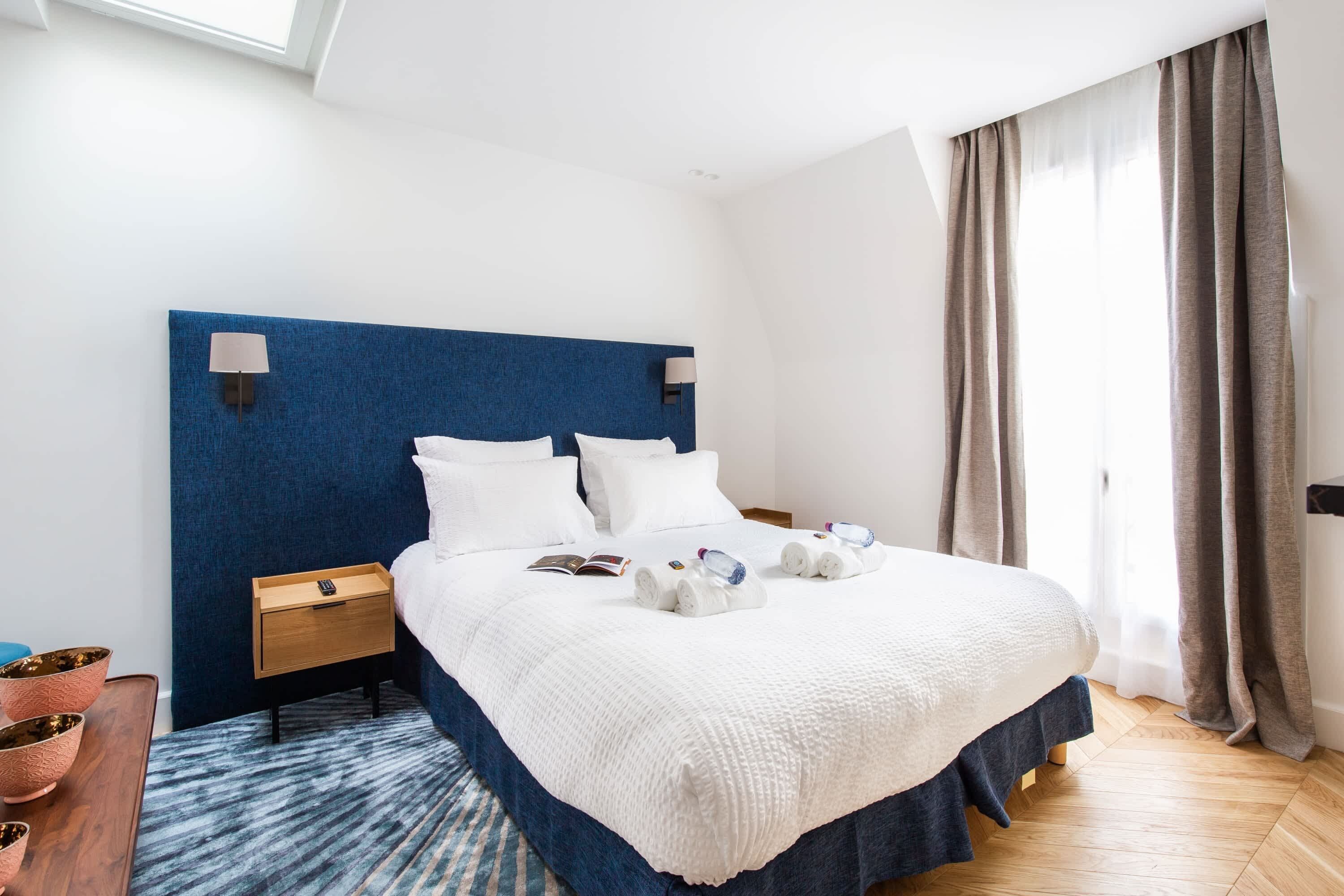 A hotel room with a large bed, a bedside table, a nightstand, a blue headboard, a window with curtains, and a wooden floor.