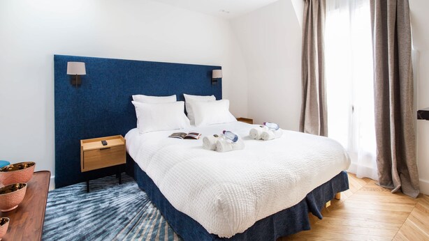 A hotel room with a large bed, a bedside table, a nightstand, a blue headboard, a window with curtains, and a wooden floor. - St Georges - Superb studio in the heart of Paris (Paris)