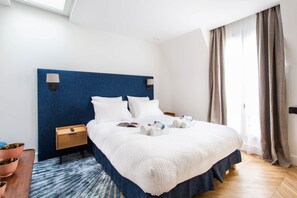 A hotel room with a large bed, a bedside table, a nightstand, a blue headboard, a window with curtains, and a wooden floor.