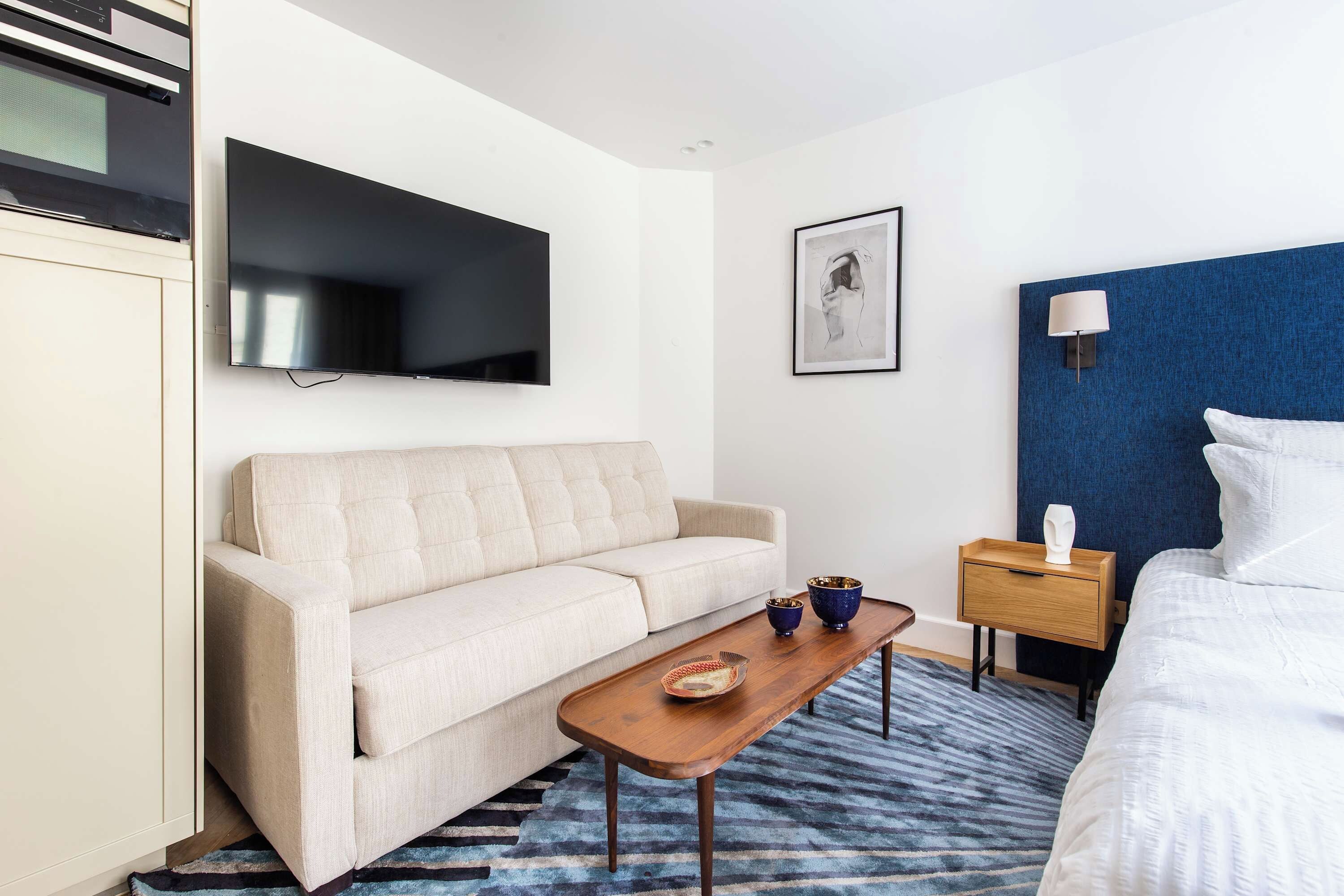 A modern hotel room with a sofa, a small wooden table, a TV, a framed artwork, a bedside table with a lamp, and a patterned rug.