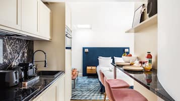 A compact kitchen with a black countertop, a built-in oven, and a sink. There is a bed with a blue headboard in the background, and a small table with a bowl of fruit.