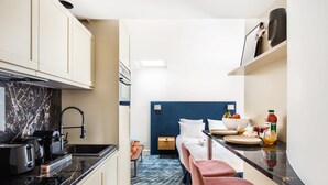 A compact kitchen with a black countertop, a built-in oven, and a sink. There is a bed with a blue headboard and a small table with a bowl of fruit. - St Georges - Exquisite studio in Paris (Paris)