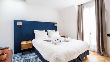 A hotel room with a large bed, a bedside table, a nightstand, a blue headboard, a window with curtains, and a wooden floor.