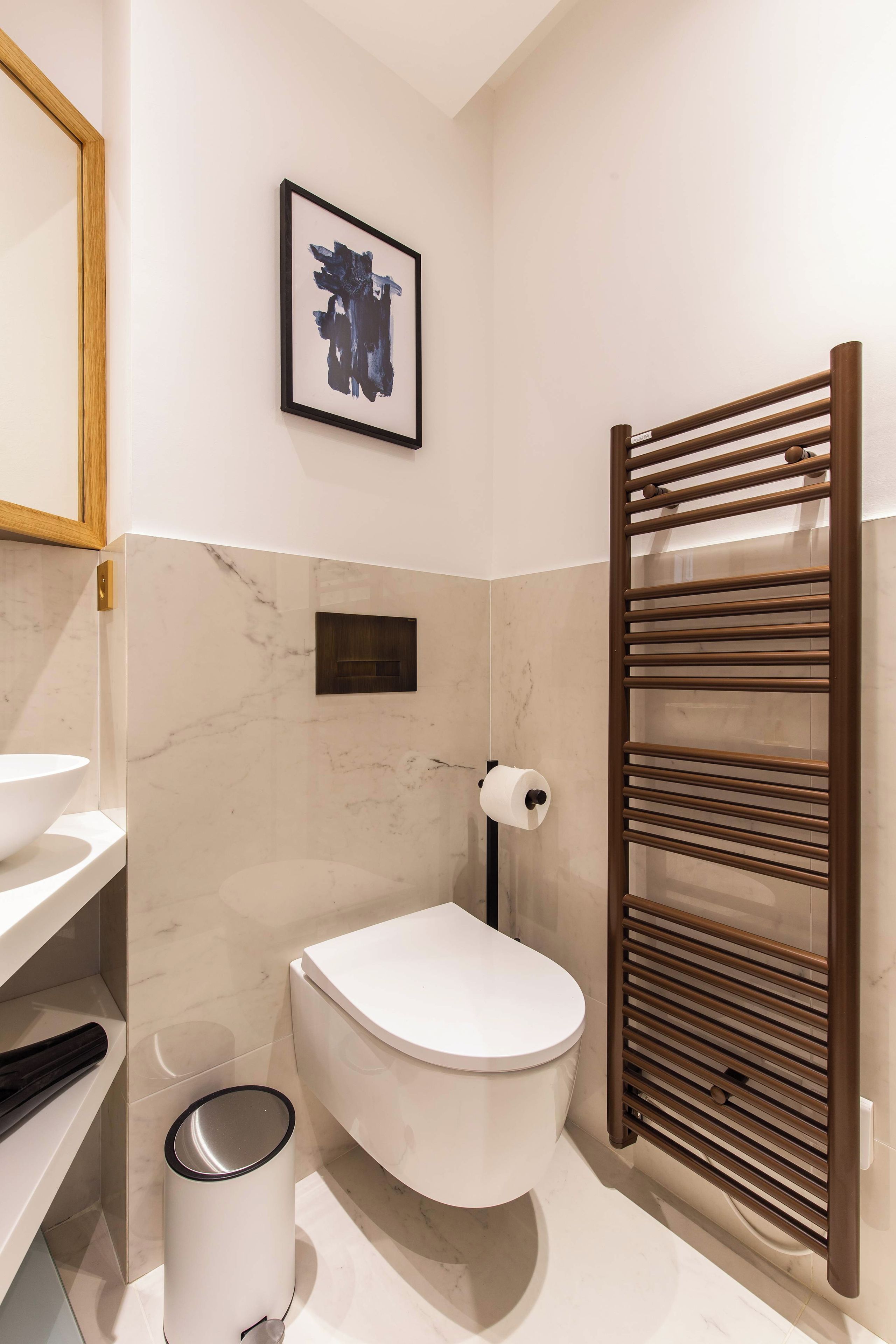 A modern bathroom with a toilet, a sink, a marble-tiled wall, a framed artwork, and a towel rack.