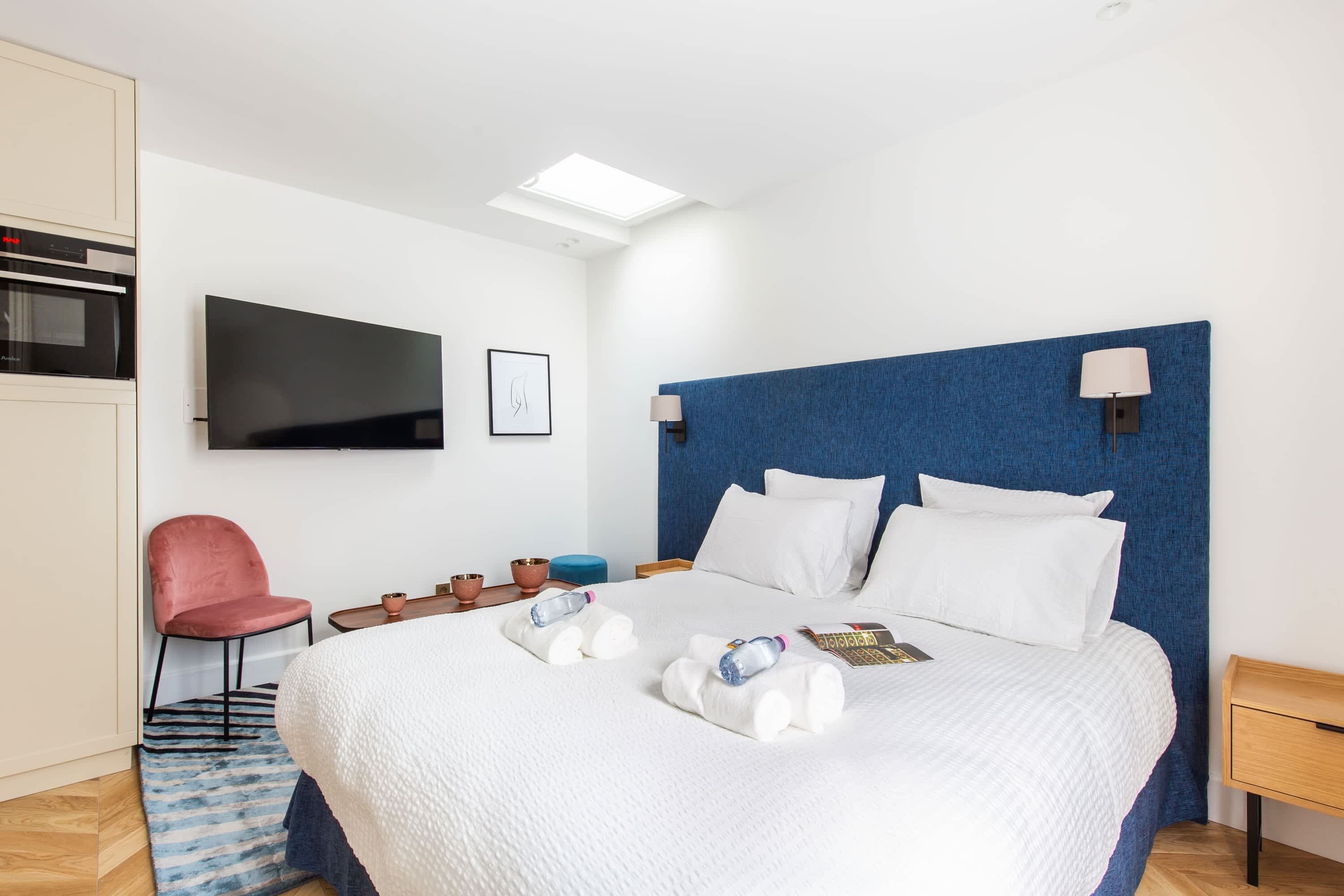 A hotel room with a bed, a TV, a chair, a small table, and a blue headboard.