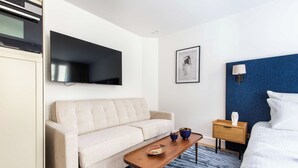 Une chambre d’hôtel moderne avec un canapé, une petite table en bois, une télévision, une œuvre d’art encadrée, une table de chevet avec une lampe et un tapis à motifs.