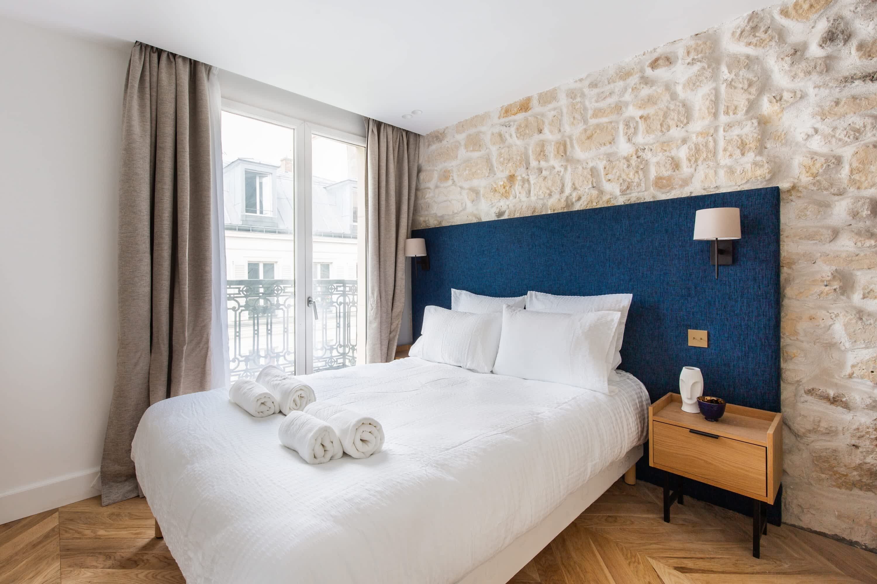 A hotel room with a stone wall, a large bed with white bedding, a wooden nightstand, and a view of the city through a window.
