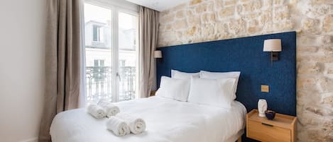 Habitación de hotel con una cama grande, una pared de piedra, una mesita de noche de madera y vistas a un balcón.
