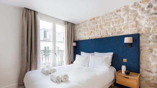 A neatly made bed with white linens and rolled towels, a wooden nightstand with a lamp, and a stone wall.