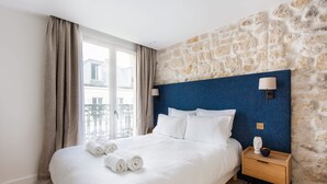 A hotel room with a stone wall, a large bed with white bedding, a wooden nightstand, and a view of the city through a window. - St Georges - One Bedroom in Paris South Pigalle (Paris)
