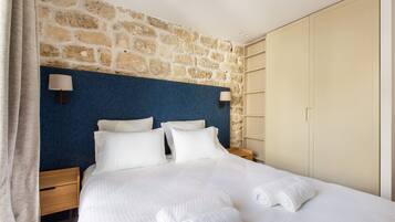 A hotel room with a stone wall, a blue headboard, a bed with white linens, two bedside lamps, and a wardrobe.