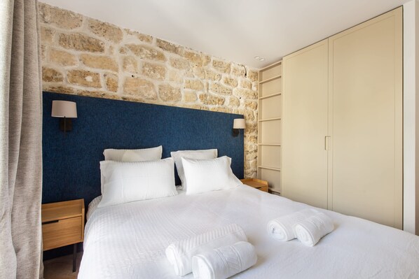 A hotel room with a stone wall, a blue headboard, a bed with white linens, two bedside lamps, and a wardrobe.